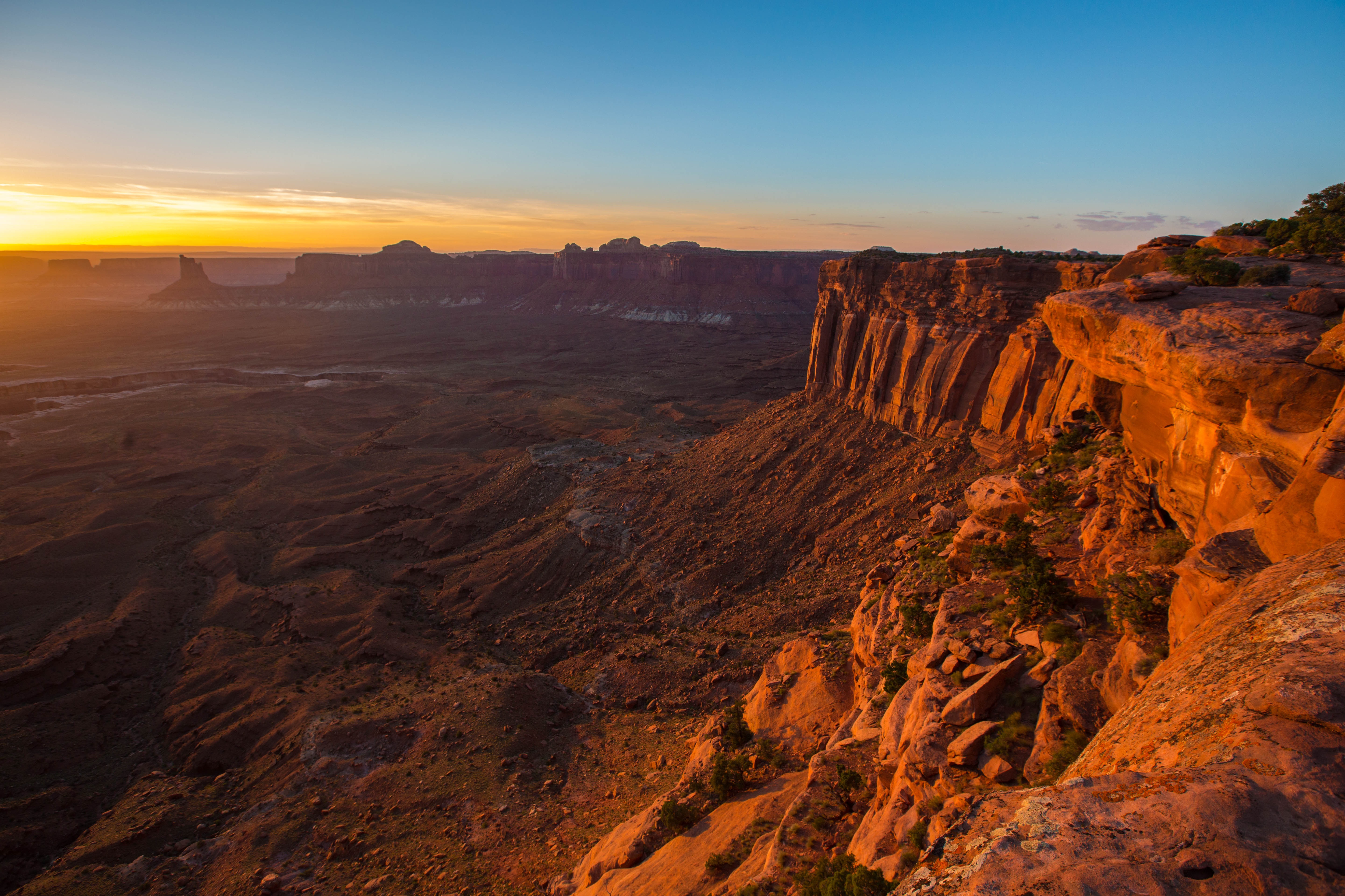 A scenic view of a canyon at sunset with rugged rock formations and a vast landscape.